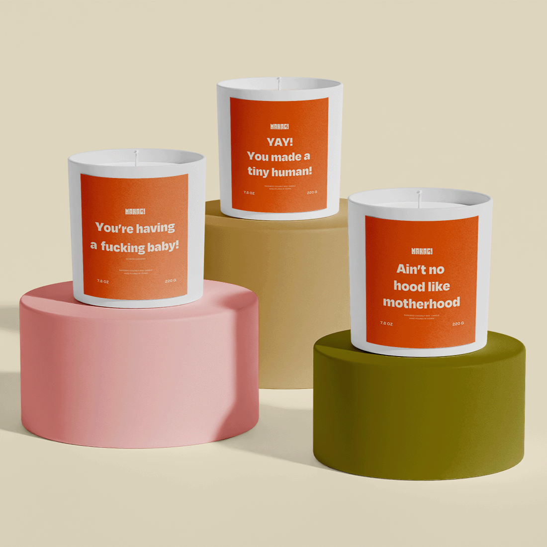 Drei Makagi-Kerzen mit humorvollen Sprüchen, darunter 'Ain't No Hood Like Motherhood', auf stilvollen geometrischen Podesten in Rosa, Oliv und Beige. Diese Kerzen sind perfekte Geschenke für werdende Mütter und feiern die Reise der Mutterschaft.