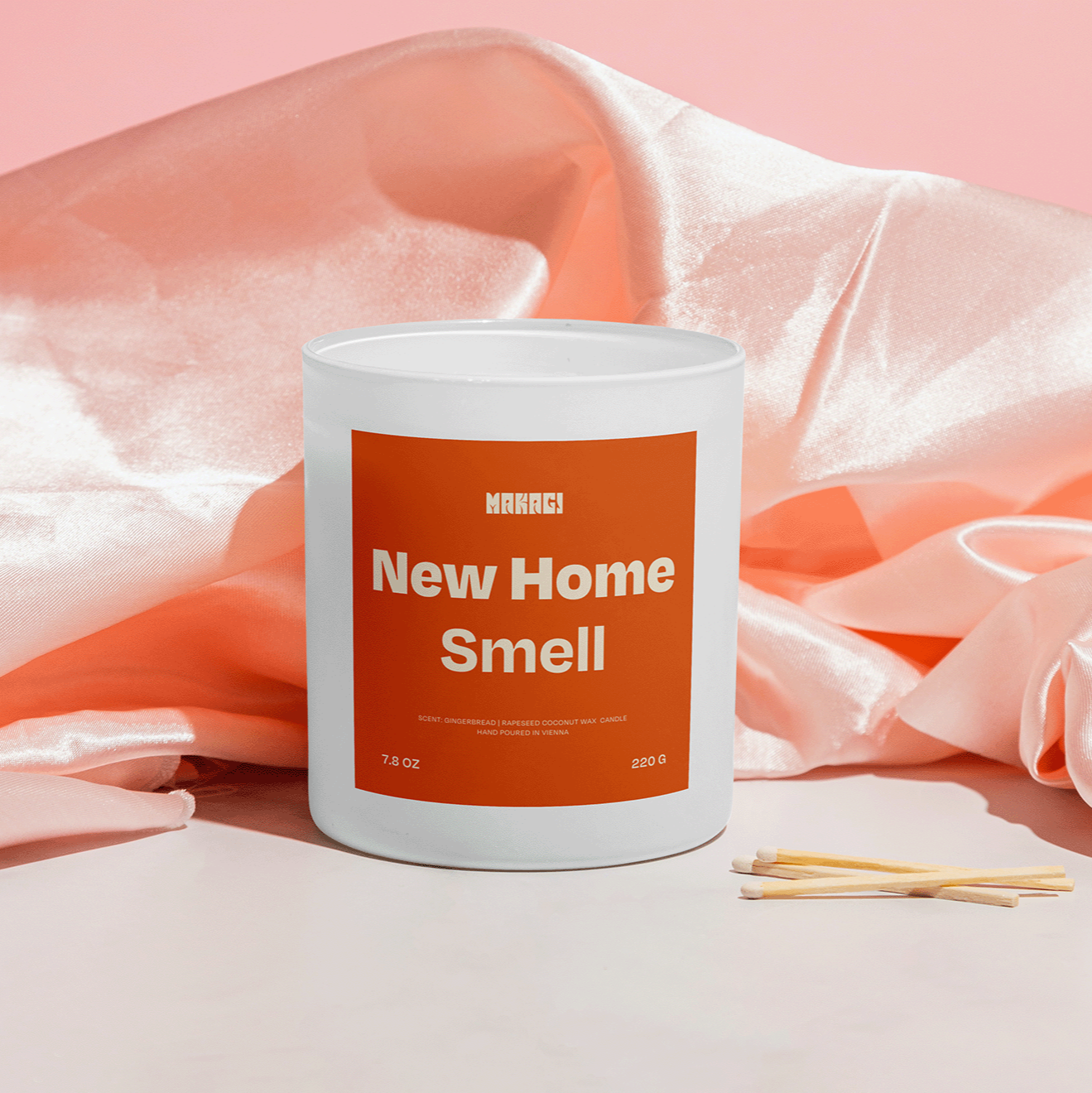 Makagi Duftkerze mit Spruch New home smell stehend auf rosa Satin Hintergrund mit Streichhölzern davor