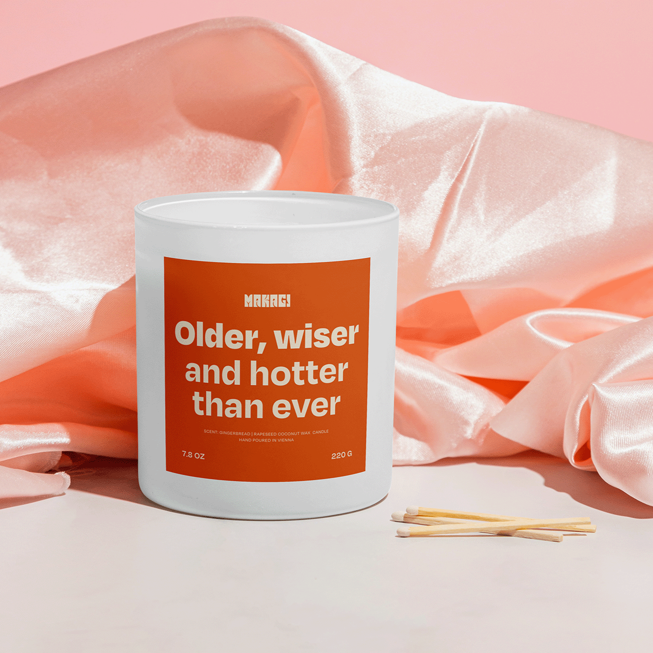 Makagi Duftkerze mit Spruch Older wiser and hotter than ever stehend auf rosa Satin Hintergrund mit Streichhölzern davor