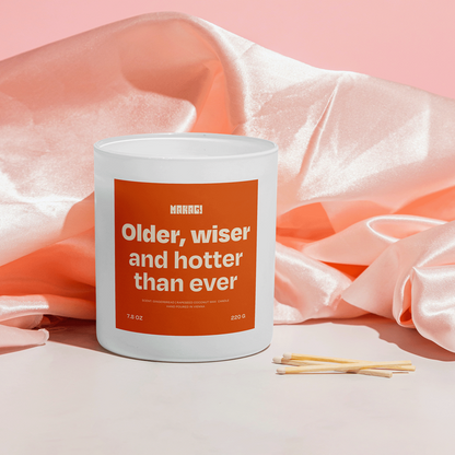 Makagi Duftkerze mit Spruch Older wiser and hotter than ever stehend auf rosa Satin Hintergrund mit Streichhölzern davor