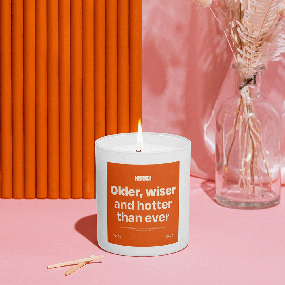 Makagi Duftkerze mit Spruch Older wiser and hotter than ever stehend auf rosa Hintergrund mit Streichhölzern davor und Vase im Hintergrund