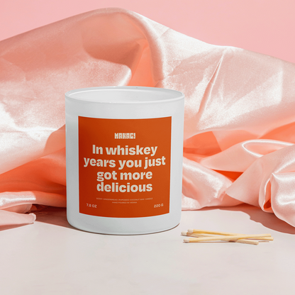 Makagi Duftkerze mit Spruch In whiskey years you just got more delicious stehend auf rosa Satin Hintergrund mit Streichhölzern davor