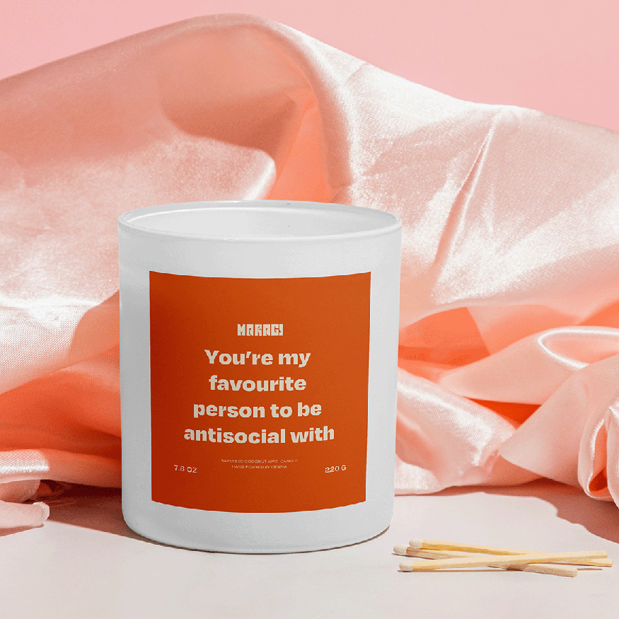 Makagi Duftkerze mit Spruch You're my favourite person to be antisocial with  stehend auf rosa Satin Hintergrund mit Streichhölzern davor