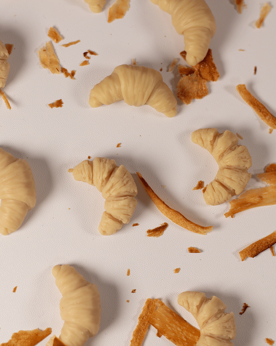 Mini-Croissant-förmige Duftwachs-Schmelzen im Duft 'Parisian Butter', kunstvoll auf einem weißen Hintergrund arrangiert, mit zerbröseltem Butterteig umgeben. Diese handgefertigten Wachsmelts verströmen den verführerischen Duft von frisch gebackenem Croissant und schaffen eine gemütliche, französische Atmosphäre.