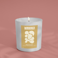 Makagi Duftkerze Isand Dream  mit dem Duft von Kokosnuss, Sandelholz und Vanille, brennend in einem eleganten weißen Glas auf einem rosa Hintergrund.
